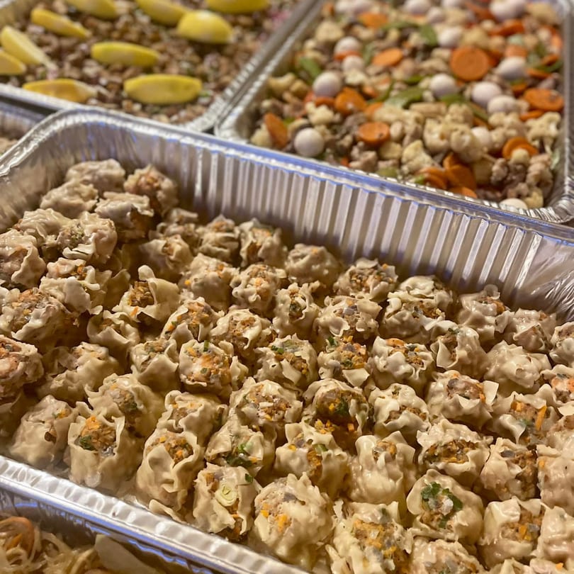 Close-up of numerous Filipino steamed siomai dumplings in a foil tray, with other dishes blurred in the background.