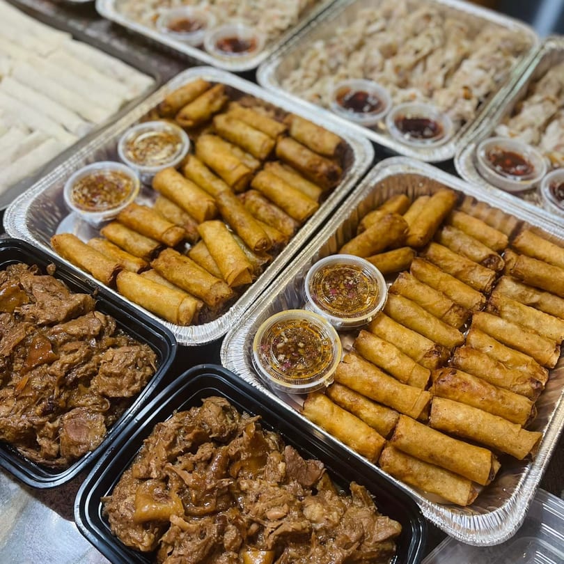 Trays of Filipino lumpia, beef adobo, and pancit, with small containers of dipping sauce.