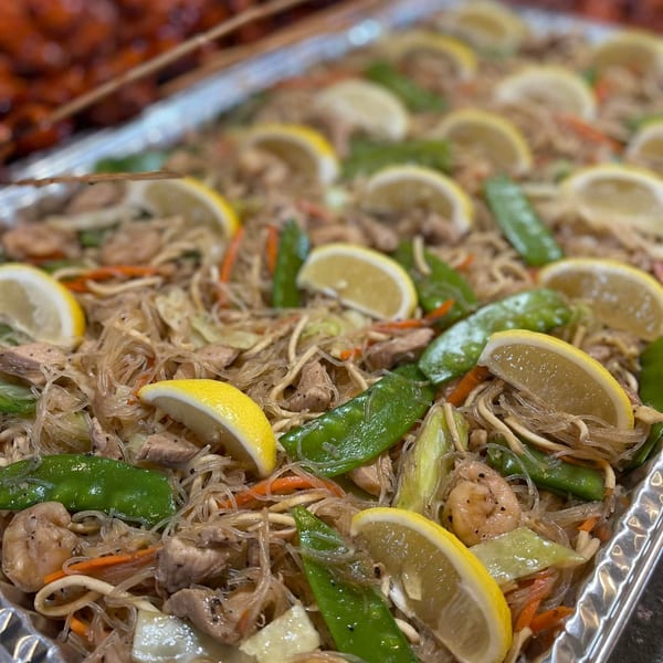 A large tray of Filipino pancit with noodles, chicken, shrimp, vegetables, and lemon slices.