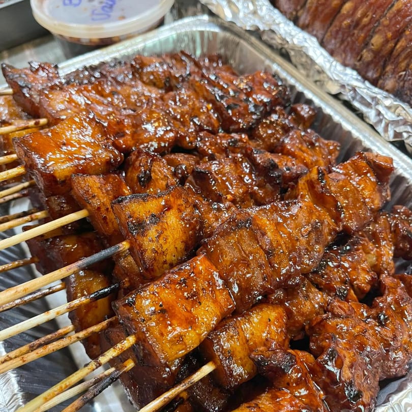 Close-up of numerous skewers of glistening, caramelized Filipino pork belly BBQ in a foil tray.